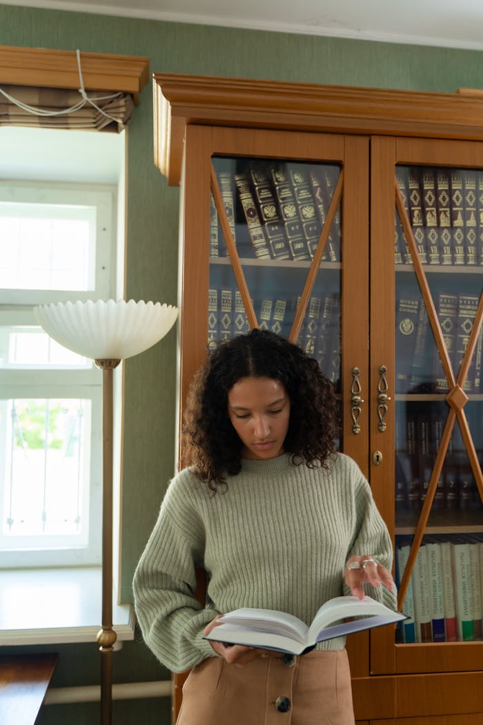 Woman reading a book in a library with wooden bookshelves, natural light filtering through.